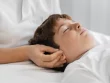 Un enfant est allongé sur le dos. Les mains de la praticienne sont posées sur le crâne de l'enfant, près de ses oreilles, durant un soin shiatsu sur un enfant.